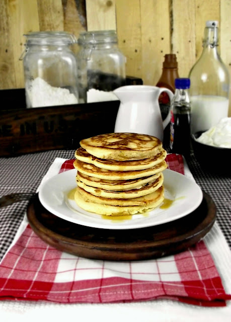 Pila de tortitas con sirope sobre un plato blanco, fondo rústico con tarros y botellas, ambiente cálido de desayuno
