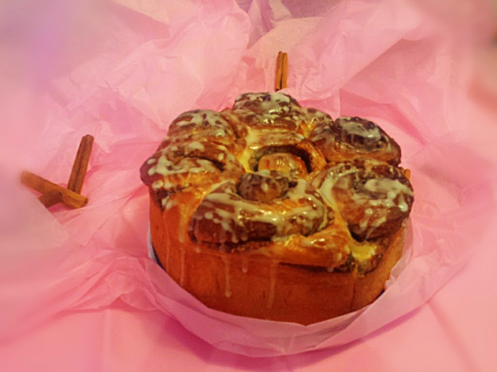 Rollito de canela glaseado con chorrito de icing sobre papel de seda rosa, con ramas de canela al lado