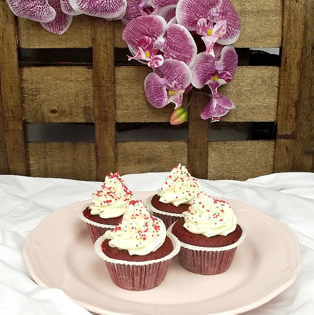 Cuatro cupcakes con frosting rojo y sprinkles sobre un plato rosa, con flores de orquídea al fondo, ambiente festivo y cálido