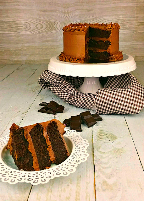 Pastel de chocolate en capas con un trozo cortado sobre un soporte blanco, trozos de chocolate oscuro esparcidos alrededor, ambiente rico y goloso