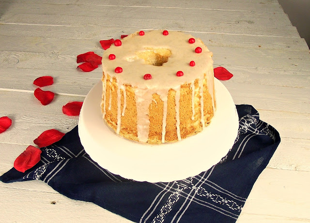 Pastel angel food cake con glaseado blanco y perlas rojas sobre fondo suave con pétalos de rosa