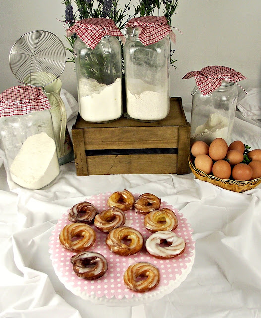 Pasteles glaseados sobre un soporte de lunares rosas, rodeados de tarros de harina y huevos, ambiente rústico de cocina casera