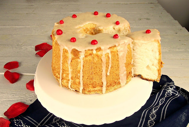 Angel food cake redondo con glaseado blanco y perlitas rojas de caramelo, un trozo cortado al lado, sobre una superficie blanca