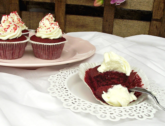 Cupcakes de terciopelo rojo con frosting blanco en remolino y sprinkles rojos sobre un plato rosa, con un cupcake cortado sobre un pañito de encaje