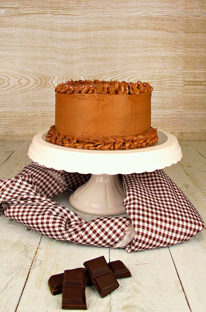 Pastel en capas cubierto de un glaseado suave color caramelo con bordes rizados, sobre un soporte blanco con decoración floral