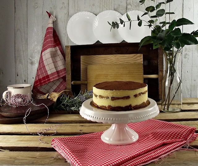 Tarta redonda en capas con crema y cobertura de cacao sobre pedestal rosa, fondo rústico con platos y vegetación